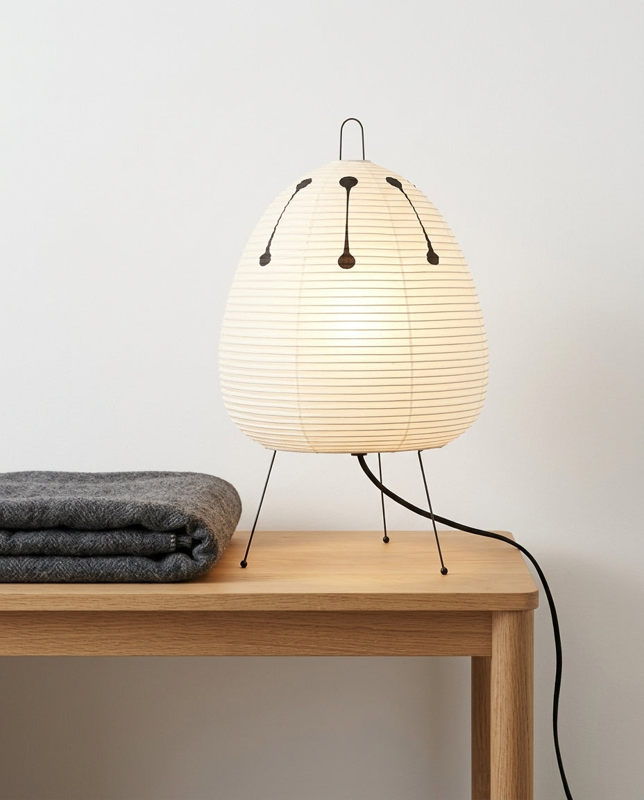 Rice Paper Tripod Table Lamp on a bedside table beside a folded throw, warm evening light