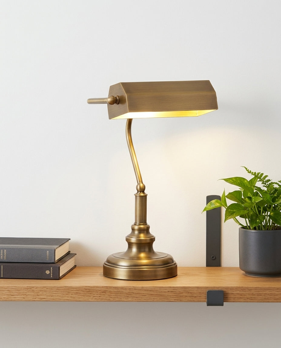 Antiqued Brass Library Desk Lamp on an oak shelf beside a few hardback books and a small plant, evening